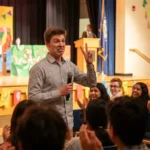 A student anchor on stage holding a microphone, laughing and making the audience laugh during a comedy anchoring segment.